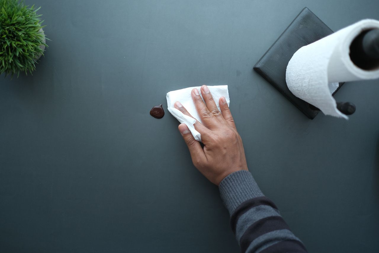 brand-01 A hand wipes a dark surface with a tissue near a paper roll and plant.